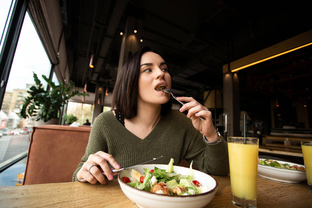 Young woman enjoys tasty meal. Attractive woman with brown hair slowly eating healthy caesar salad and looking away. Glass of orange juice. Restaurant or cafe on background. Cozy atmosphere. - Foto, afbeelding