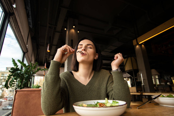 満足した若い女性は、彼女の口の中にフォークを入れて、彼女の目を閉じる、レストランでおいしいサラダを楽しんでいます。良い顧客サービス。健康的な食事の概念。闘わずに減量. - 写真・画像