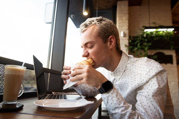 Blond man enjoying croissant in cafe. Coffee shop on background. Lunch time concept. Remote work. Yummy croissant. Cup of latte and modern laptop on table. Young attractive man biting bake. - Zdjęcie, obraz
