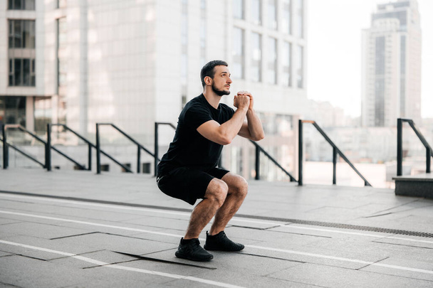 Fit man training air squat exercise. Fit male exercising crossfit outside. Young handsome caucasian male fitness model and instructor outdoors. Black sportswear. Sport in big city concept. - Foto, immagini