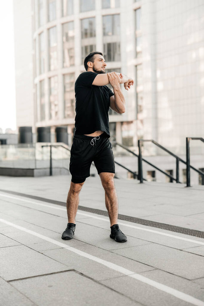 Sporty man stretching arms before jogging. Runner in black sportswear excercising at morning. Healthy lifestyle concept. Morning city on background. Active living. Outdoor fitness. - Foto, imagen