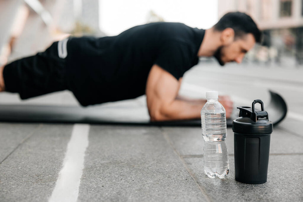 Concentrated man doing plank position while exercising on black yoga mat. Water bottles lying near. Blurred man in black sportswear exercising outdoors. Morning warm up. Side view. - Photo, Image