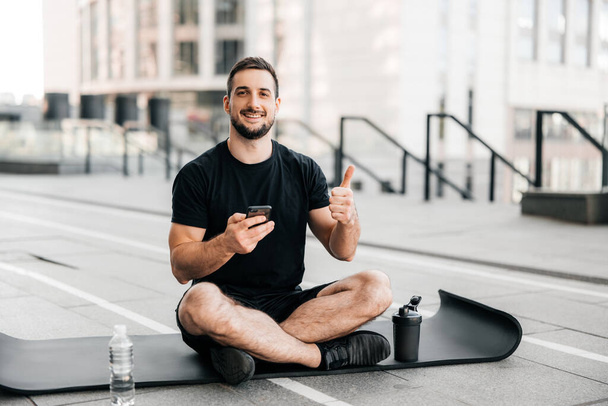 Man resting after outdoors excercising with smartphone in hand and thumbing up. Sports water bottle on black yoga mat. Sporty man after practicing yoga, relaxing on mat, texting on phone. - Photo, Image