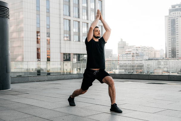 Athletic man practice Low Lunge pose with raised arms. Young man in black sports t-shirt and leggins stretching in yoga pose. Urban sport concept. Morning warm up. - Photo, Image