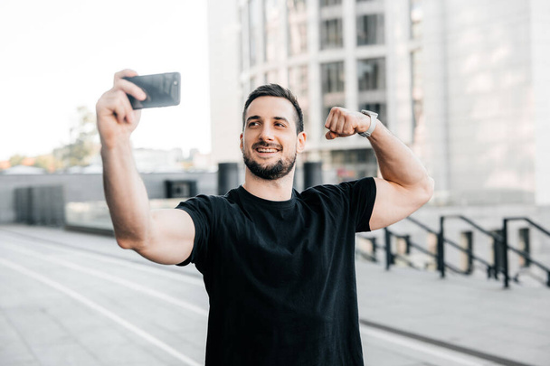 Attractive man shows biceps and takes a selfie. Social media concept. Male athlete in black t-shirt shows to friends on a videocall his fit body. Morning workout. Urban sport concept. - Photo, image