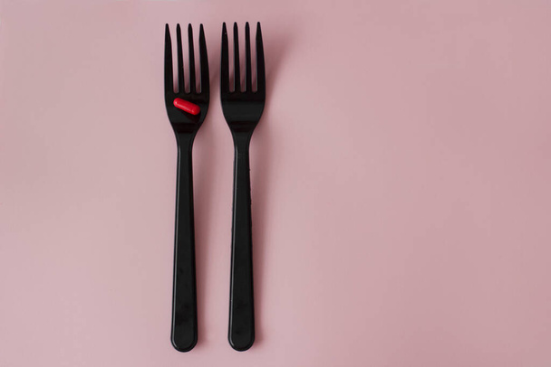 A red pill on a black fork. Two black forks with a red capsule on a pink background. - Фото, изображение
