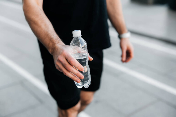 Bebe un poco de agua Mans mano sostiene una botella de plástico de agua. Asfalto gris sobre fondo. Hombre en ropa deportiva negra corriendo. Tiempo de hidratación Entrenamiento afuera. - Foto, imagen