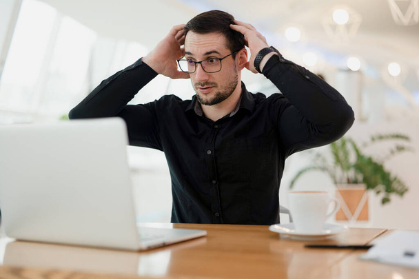 Whats happening Shocked smart man in eyeglasses looking on his laptop and holding head by his hands. Error in the program. Male programmer is shocked by how many bugs there are in the code. - Foto, Imagen