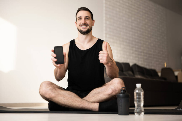 Satisfied man thumbs up and shows favorite sports mobile app. Workout everyday concept. Two sports water bottle. Happy man sits on black yoga mat with crossing legs. Smart lifestyle concept. - Photo, Image