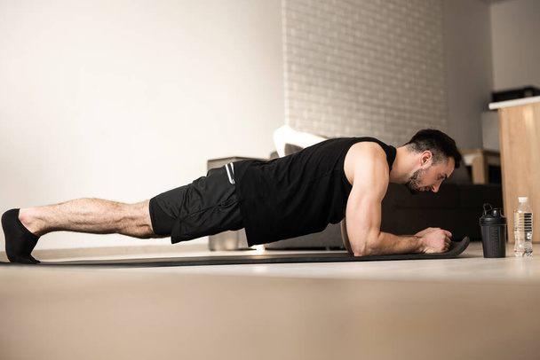 Plank it Training at home. Confident young man wearing black sportswear and doing plank position while exercising on the floor in light loft interior. Hard workout. - Photo, Image