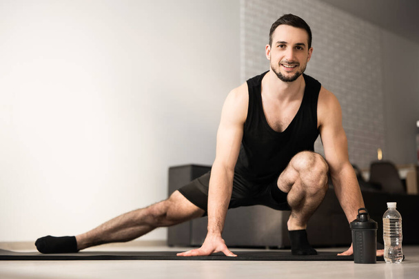 Sporty man doing side lunge exercise. Male athlete with fit body warming up, doing leg stretch workout at home. Take care of yourself Modern light room on background. Black sportswear. - Photo, Image