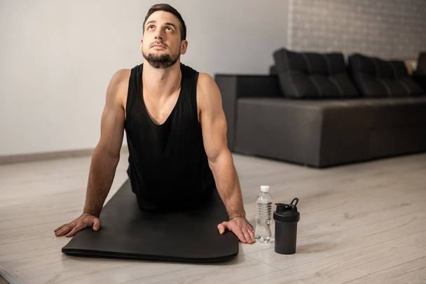Fitn man meditating while doing cobra pose in living room. Indoor shot of handsome young man practicing yoga. Morning workout. Black sportswear. Plastic water bottle. Strong arms. - Photo, Image