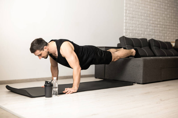 Sporty man pushing from floor with legs on sofa. Workout on black yoga mat. Man in black sportswear exercising at home. White modern room on background. Strong man excercising to have a fit body. - Valokuva, kuva