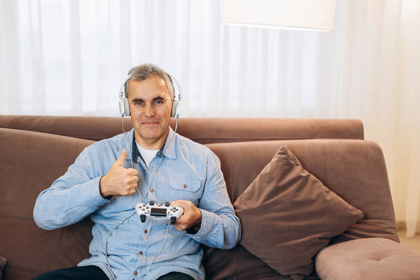 Volwassen gamer man spelen video game met behulp van joystick en hoofdtelefoon blij met grote glimlach, duimen omhoog. Uitstekend teken. Woonkamer op achtergrond. Man in casual kleding. - Foto, afbeelding