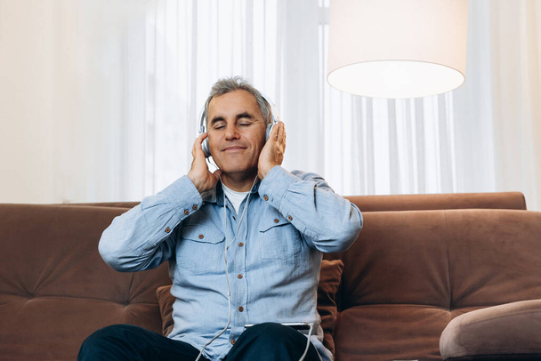 Großartige Musik Mann mittleren Alters sitzt auf einem braunen Sofa, hält Kopfhörer in den Händen und genießt seine Lieblingsmusik. Gemütliches Wohnzimmer im Hintergrund. Mann in Freizeitkleidung nutzt Smartphone und lächelt. - Foto, Bild