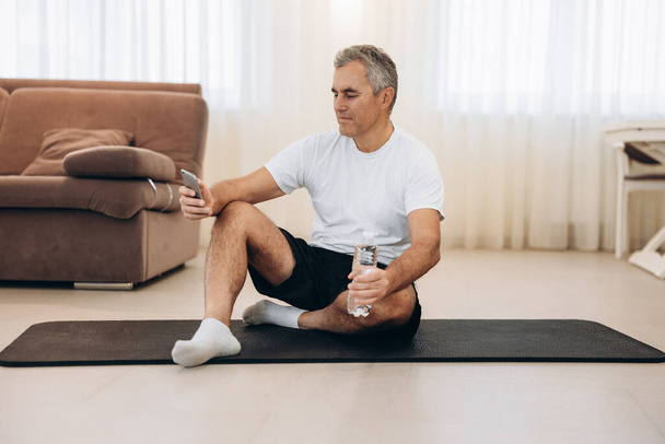 Legen Sie Ihr Handy ab, wenn Sie während des Trainings einen starken Körper wollen. Älterer Mann sitzt auf Yogamatte und schreibt SMS mit dem Smartphone. Alt mann takes ein rest nach schwer training. - Foto, Bild