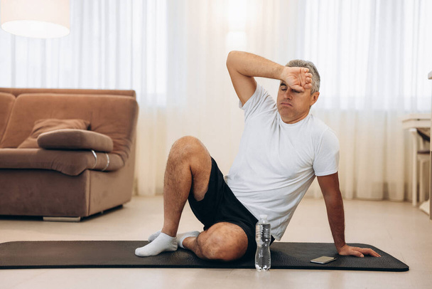 Senior man wipes sweat of his forehead after hard home workout. Healthy lifestyle concept. Living room on background. Man tired after intensive training. Plastic water bottle near black yoga mat. - Photo, Image