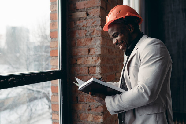 African American man in a construction helmet looks out of the window at the building and writes in a notebook - Zdjęcie, obraz