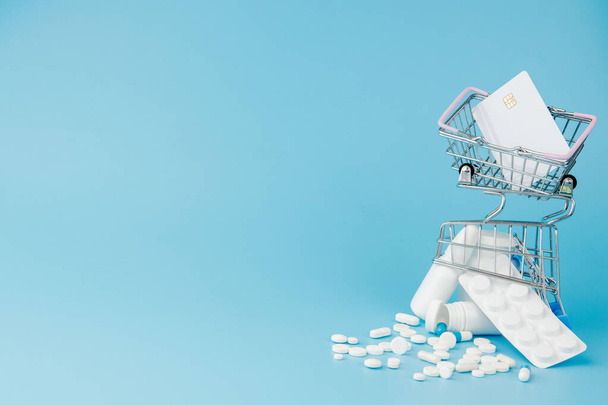 scattered variety pills, drugs, spay, bottles, thermometer, syringe and empty shopping trolley cart on blue background. pharmacy shopping concept. - Photo, Image