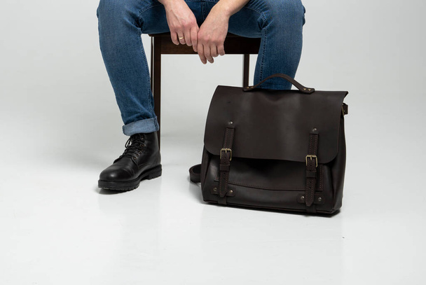 Man in a blue jeans and black boots sits on a chair with a brown mens shoulder leather bag for a documents and laptop on a white floor. Mens leather satchel, messenger bags, handmade briefcase. - Photo, Image