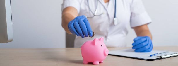 A faceless female doctor puts a coin in a piggy bank. Widescreen - Photo, Image