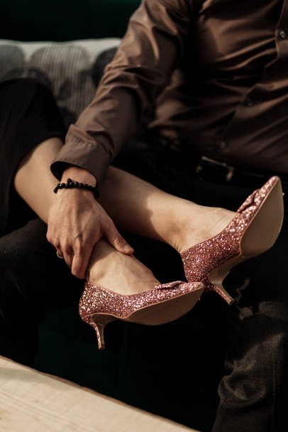 Close up a woman putting her legs in rose golden high heels on man. Man holding womans feets on his knees. Elegant couple sitting on sofa. Valentines love day - Photo, Image