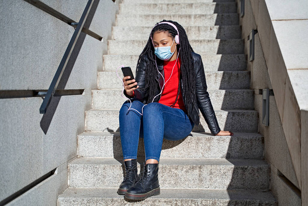 Een portret van een Afro-Amerikaanse vrouw met een gezichtsmasker met een mobiele telefoon luistert naar muziek in de straten van de stad.Het haar is gevlochten tot dreadlocks. Begrip nieuwe normaliteit - Foto, afbeelding