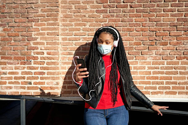 Un retrato de una mujer afroamericana en una máscara facial con un teléfono celular está escuchando música en las calles de la ciudad.El pelo está trenzado en rastas. Concepto de nueva normalidad - Foto, Imagen