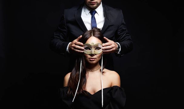 la mujer hermosa en la máscara en las manos del hombre en el traje. Pareja sobre fondo negro - Foto, Imagen