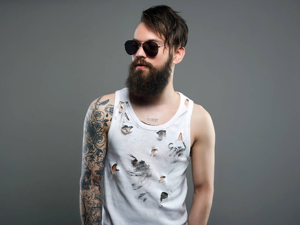 handsome man in sunglasses. Brutal bearded hipster boy in glasses and vintage torn dirty shirt. beard, haircut, tattoo - Photo, Image