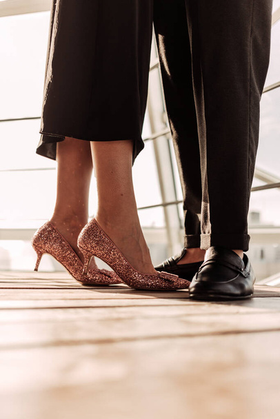 Nahaufnahme einer Frau in elegantem schwarzen Kleid und roségoldenen High Heels, die mit ihrem Mann in schwarzen Hosen und Schuhen zusammenbleibt. Elegantes Paar vor Sonnenlicht auf Holzboden in der Nähe von Geländern - Foto, Bild