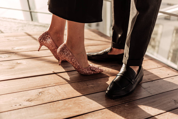 Close up a woman in elegant black dress and rose golden high heels staying together with her man in black pants and shoes. Elegant couple staying in front of sunlight on wooden floor near railings - Photo, Image