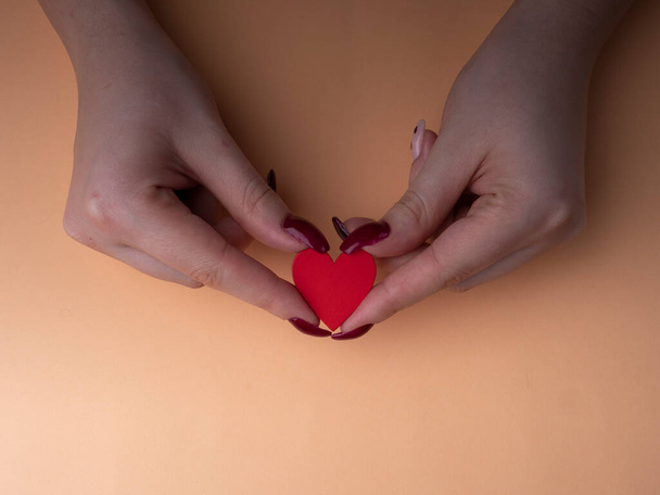 Κομψό γυναικείο μανικιούρ. Νεαρά θηλυκά χέρια κατέχουν ένα κόκκινο σχήμα καρδιάς, καρφιά με κόκκινο μανικιούρ, σε πορτοκαλί φόντο. Μανικιούρ στο σπίτι κατά τη διάρκεια πανδημίας - Φωτογραφία, εικόνα