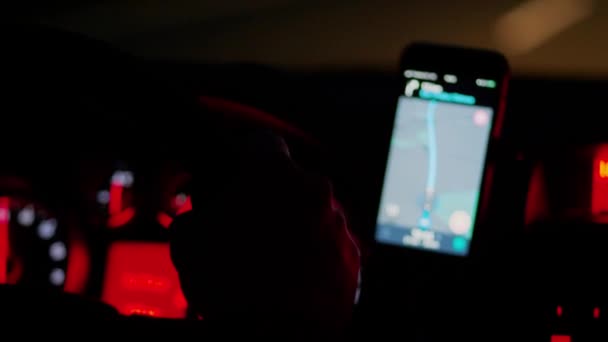Dentro del coche de conducción por la noche, teléfono móvil desenfocado con iluminación de panel de navegación y control rojo, enfoque en la mano sosteniendo el volante, primer plano de mano. Persona libre por guía de ruta en el camino de la tarde. Movimiento de transporte borroso en la oscuridad - Imágenes, Vídeo