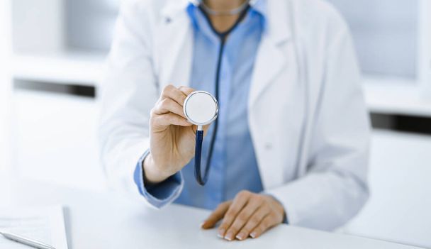 Unknown woman-doctor in blue blouse is holding stethoscope head while sitting at the desk in clinic, close-up. Physician ready to examine and help patient. Medicine concept - Foto, afbeelding