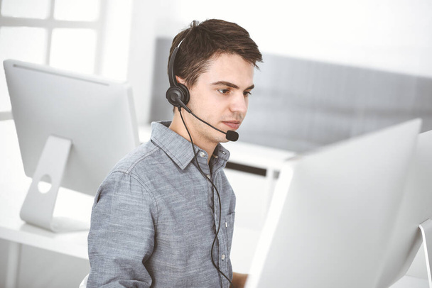Casual dressed young man using headset and computer while talking with customers online. Call center, business concept - Фото, изображение