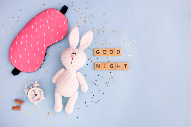 Healthy sleep concept. Amigurumi hare with alarm clock and sleep mask on a blue background with confetti stars. Top view, flat lay, copy space. - Foto, imagen