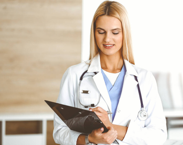 Doctor woman controls medication history records and exam results while using clipboard in sunny hospital . Medicine and healthcare concept - Photo, image