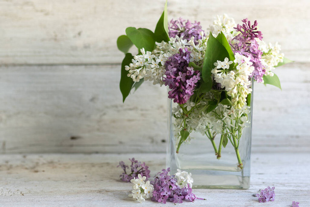 Un ramo de lilas primaverales de color púrpura y blanco en un jarrón sobre un fondo de madera clara. Recipiente transparente de vidrio con agua. Composición aromática de disposición horizontal. Enfoque suave - Foto, Imagen
