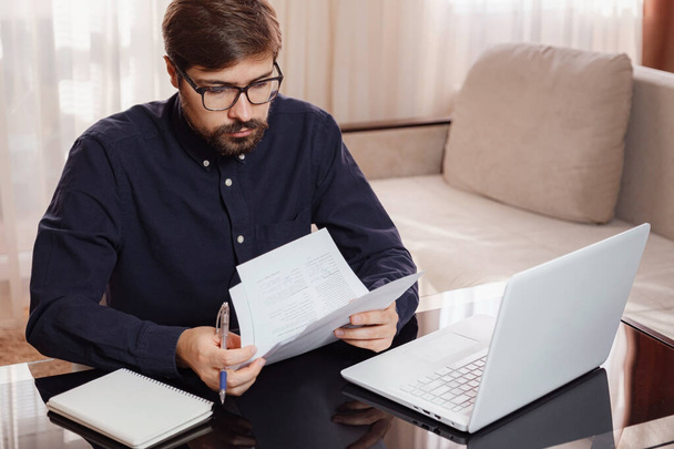 Entrepreneur in eyeglasses works with a laptop and keeps a document in a home office. Man holding paper documents, chatting online with clients on laptop at workplace. - Valokuva, kuva