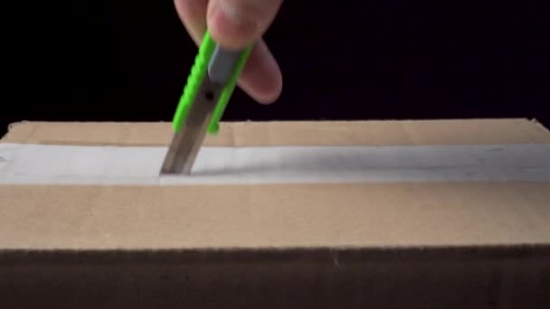 Desempaquetar una caja de cartón de paquete de correo con un cortador de cerca. Cortar la cinta de embalaje. Unboxing sobre fondo negro - Metraje, vídeo
