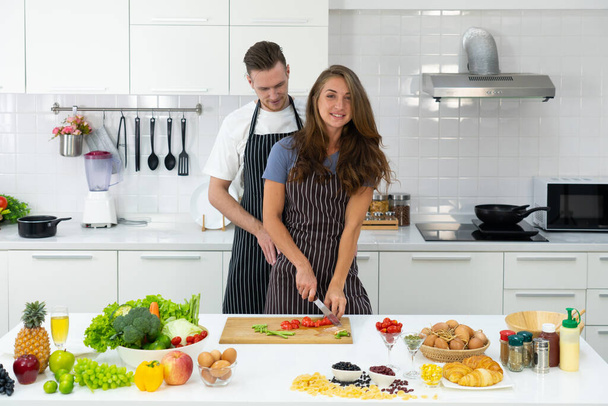egy fiatal nő zöldséget aprít, hogy egészséges salátát főzzön, míg a férj megöleli a konyhában. pár együtt koncepció - Fotó, kép