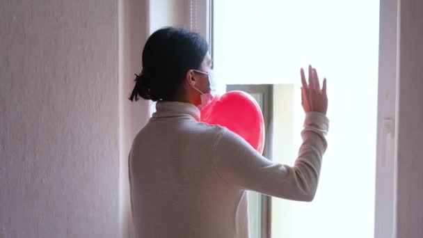 Szomorú lány egy védő orvosi maszkban, piros lufival az ablak mellett.. - Felvétel, videó