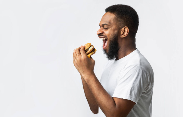 Вид збоку голодний чорний чоловік їсть бургер, білий фон
 - Фото, зображення