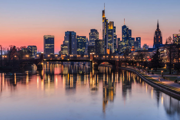 Frankfurt skyline in the evening at sunset. High-rise buildings from the financial district and river Main with reflections. Illuminated houses and bridge - Фото, изображение