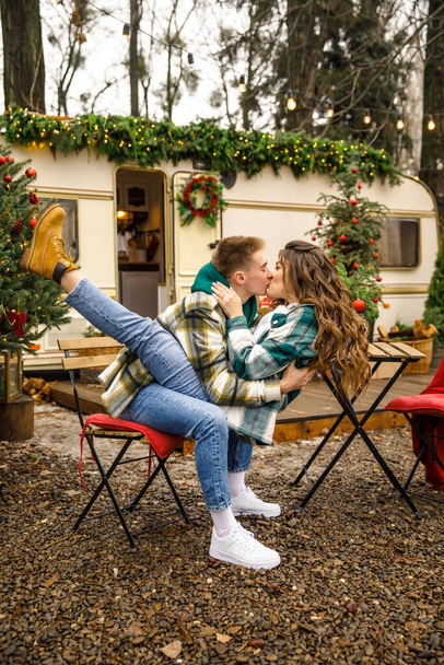 young beautiful couple dressed in a green jacket in a cage and blue jeans, sitting at a table against the background of a trailer with Christmas decorations - Fotó, kép