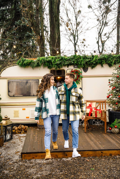 young beautiful couple dressed in a green jacket in a cage and blue jeans are walking in the park against the background of a trailer with Christmas decorations - Φωτογραφία, εικόνα