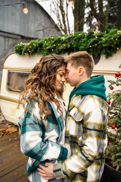 young beautiful couple dressed in a green jacket in a cage and blue jeans are walking in the park against the background of a trailer with Christmas decorations - Foto, Imagen