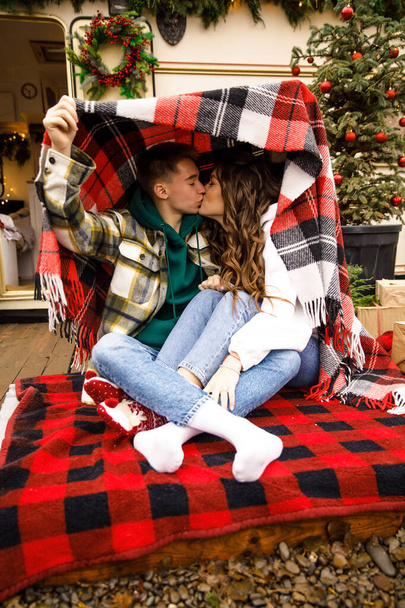young beautiful couple dressed in a green jacket in a cage and blue jeans, sit on a red blanket on the background of a trailer with Christmas decorations - Foto, Bild