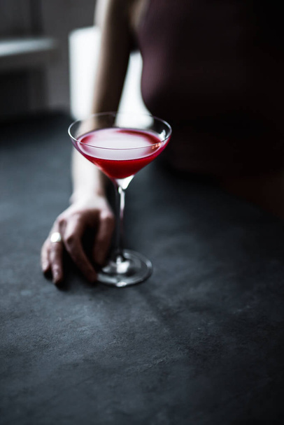 Kırmızı bir ahududu kokteyli güzel bir coupe bardağında kızın ellerinde, arka ışık, karanlık ve karamsar bir fotoğraf. - Fotoğraf, Görsel